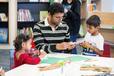 A father and his two children, all of whom are Brown, are sitting around a table in a Library, taking part in arts and crafts activities.