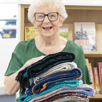 An elderly lady holds a pile of knitted scarves
