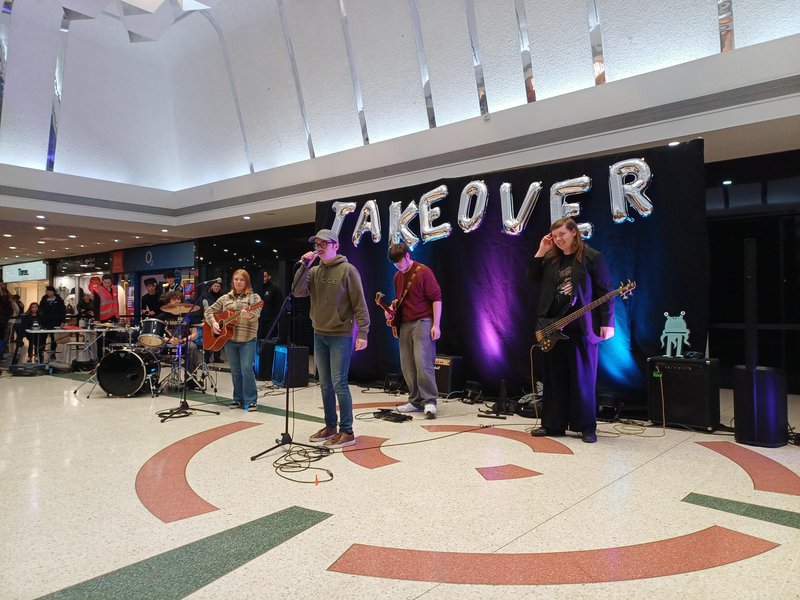 A band performing in a shopping centre. There are 5 members, a singer in the centre, a bass guitarist 2 guitarists and a drummer. There is a balloon sign that says Takeover behind them.