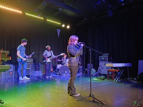 An image of a band performing. The lead singer is standing at the front with a microphone, there is a guitarist, bassist and drummer playing behind.
