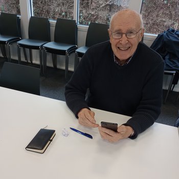 A bald white man in his sixties is smiling at the camera whilst holding a smartphone.