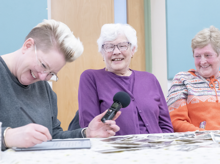 Three women sat at a table laughing, one holding a microphone